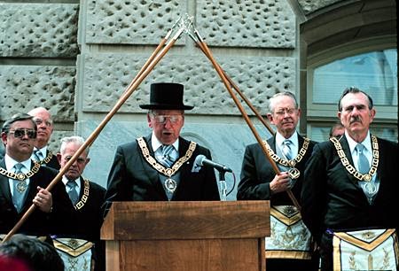 A Mason, surrounded by five brothers, speaks at a podium in traditional dress at a Masonic Cornerstone Laying Ceremony