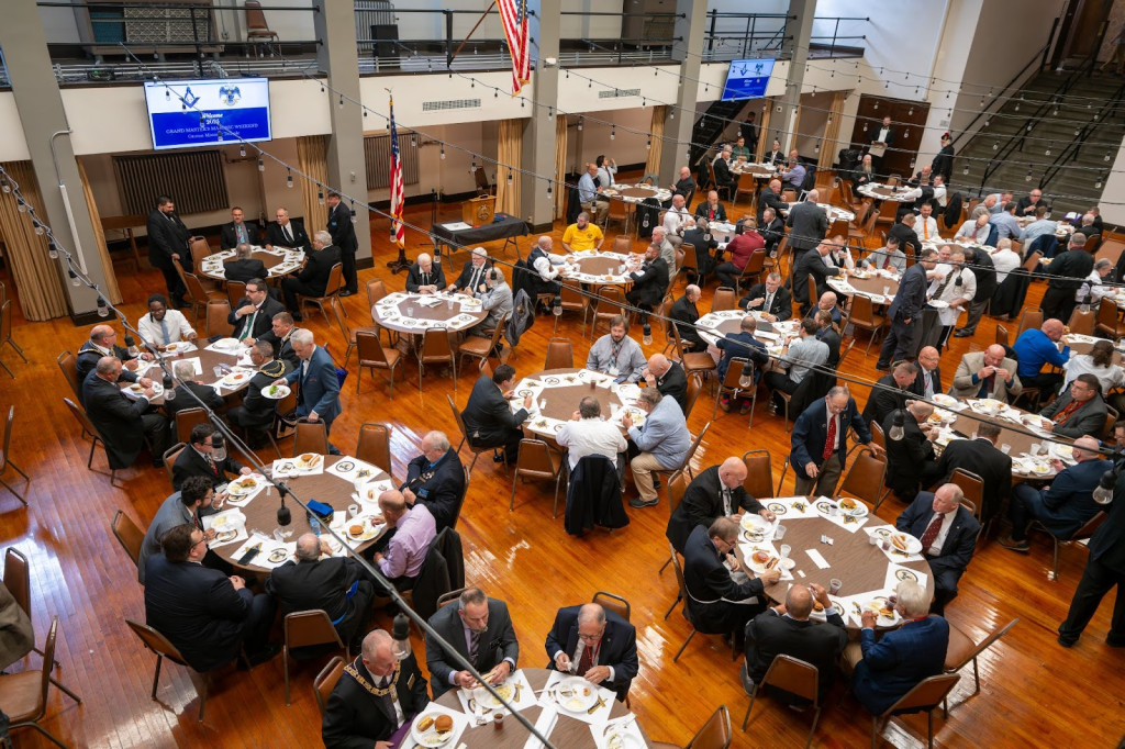 An overhead shot of the function. Tables with men seated at each, eating food, birds eye view of the event.