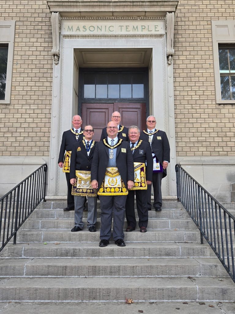 Six smiling men (all past and present Grand Master's) standing in a V shape on a grey stairway, all wearing Masonic Apron's and their formal Grand Master attire.