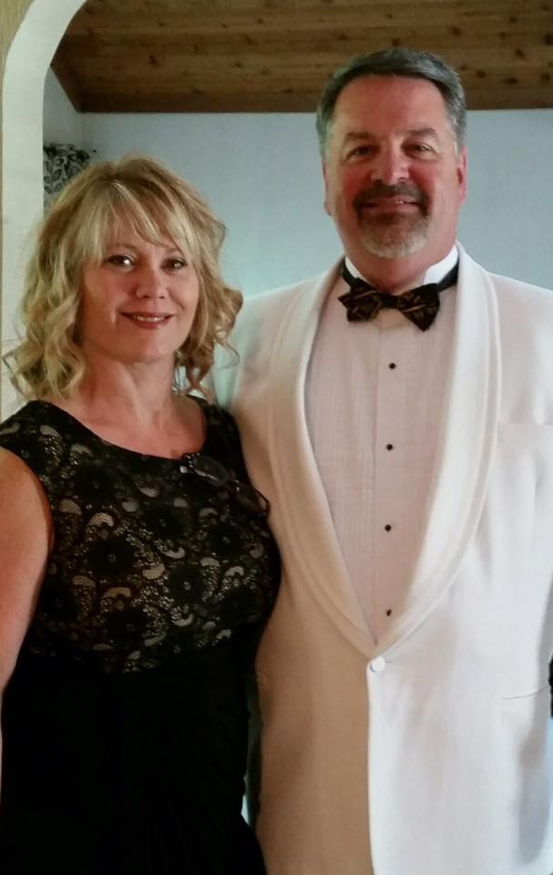 A man in a white tuxedo and bow tie standing beside a woman in a black lace evening gown, both smiling at a formal event.
