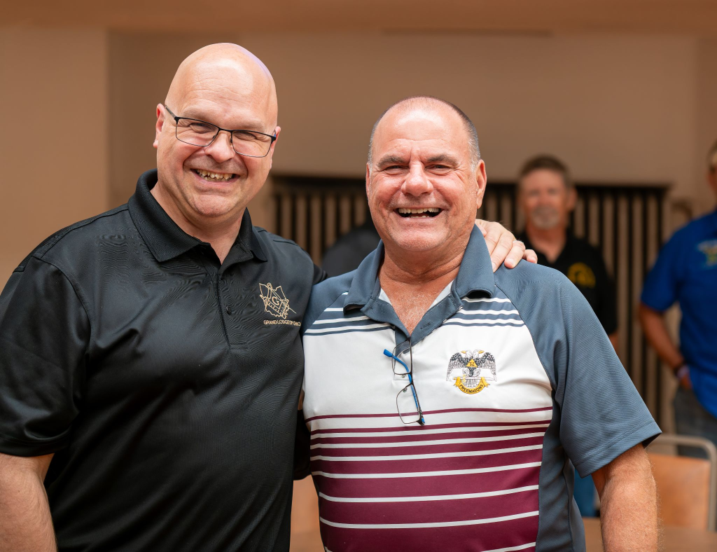 Two men smiling next to each other who are both Masons, Grand Master Bill Carter on the left wearing a black polo, and a fellow Mason on the right wearing a striped polo.