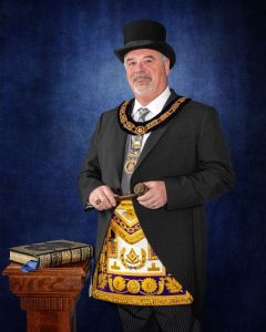 Portrait of Most Worshipful Brother Shawn Johnson, Grand Master of the Grand Lodge of Ohio, dressed in formal Masonic regalia with apron and collar, holding a gavel beside a Masonic Bible on a wooden pedestal.