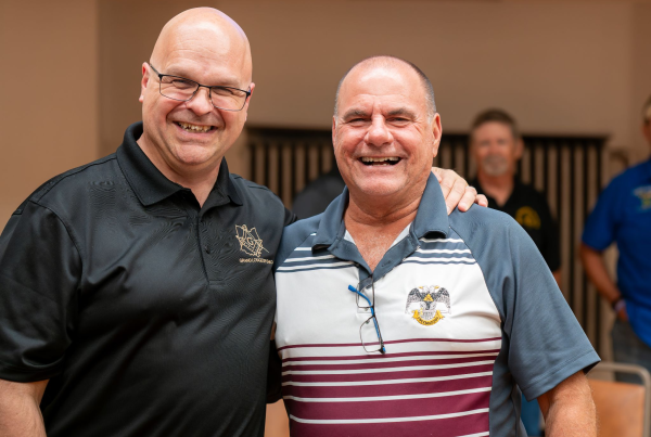 Two men smiling next to each other who are both Masons, Grand Master Bill Carter on the left wearing a black polo, and a fellow Mason on the right wearing a striped polo.