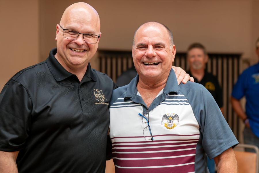 Two men smiling next to each other who are both Masons, Grand Master Bill Carter on the left wearing a black polo, and a fellow Mason on the right wearing a striped polo.