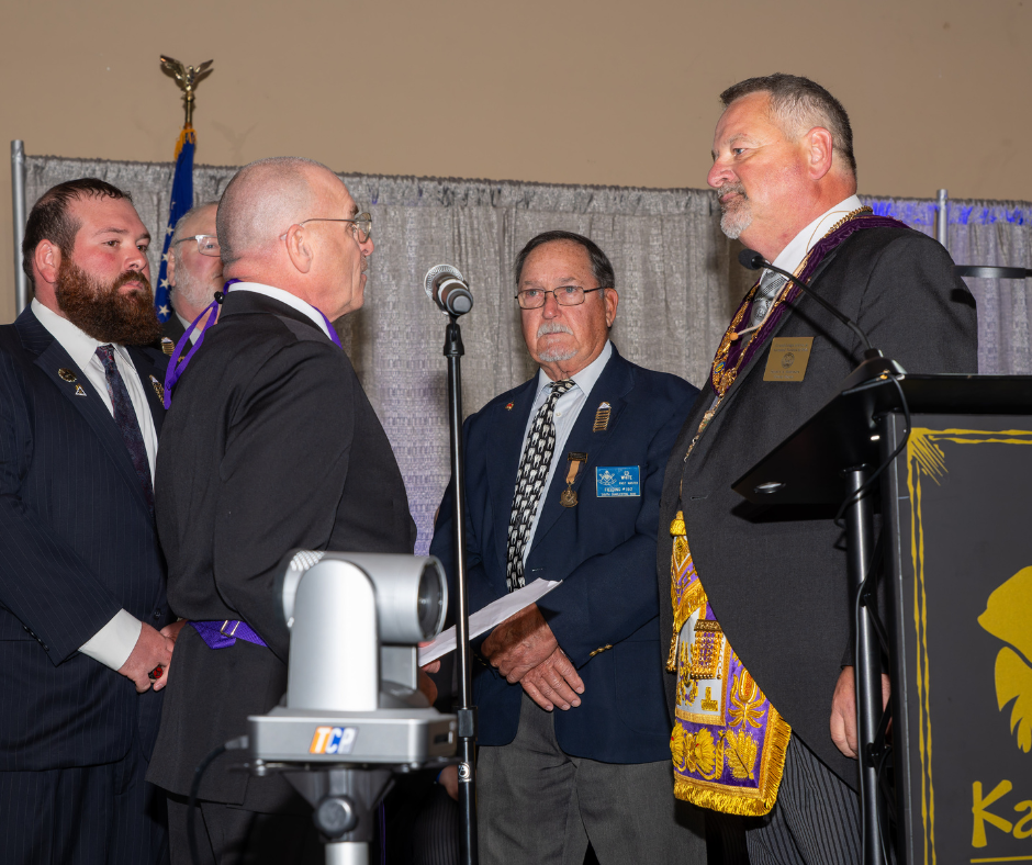 Grand Master Shawn Johnson participates in his installation ceremony, standing at a podium across from several Masonic leaders in formal attire, with an American flag and gathered attendees behind them.