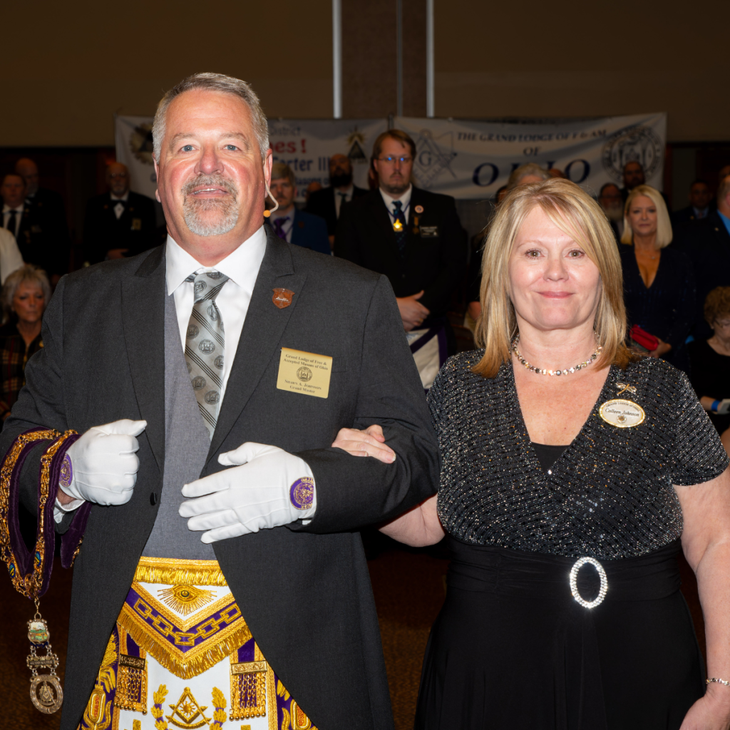 Grand Master Shawn Johnson stands arm-in-arm with First Lady Colleen Johnson during an Ohio Freemasonry event, with attendees seated and standing in the background.