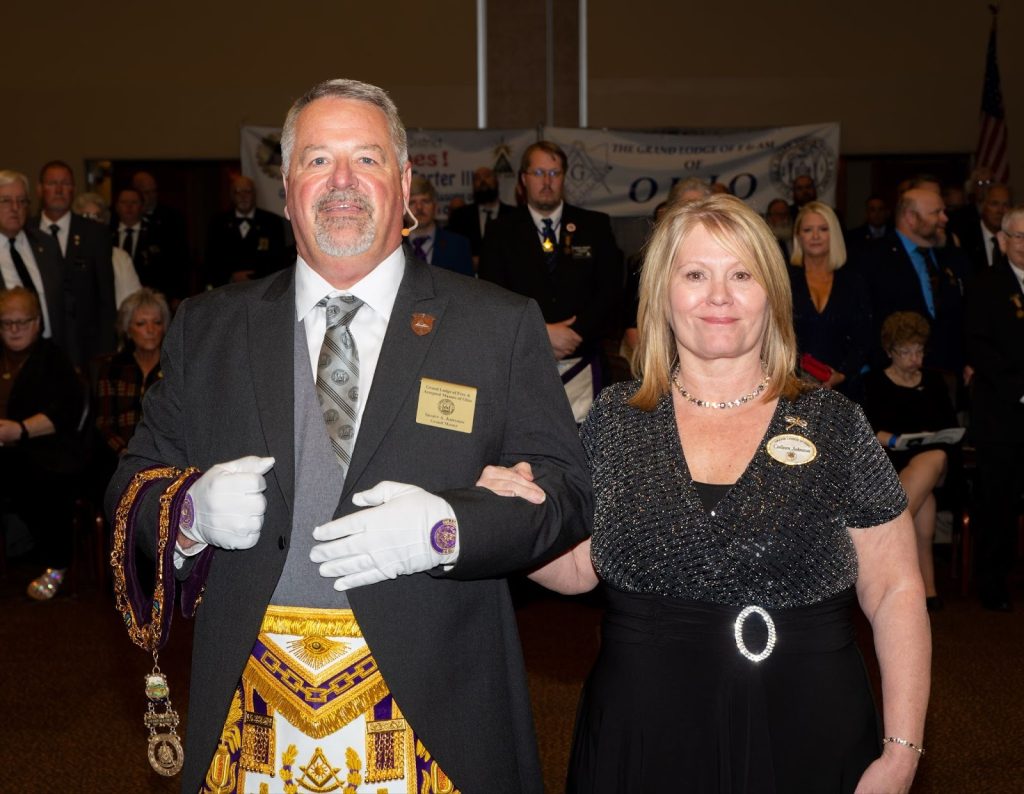 A couple stands arm-in-arm in formal attire in a crowded ceremonial hall, surrounded by Masons in suits and regalia during the 2025-2026 installation ceremony.