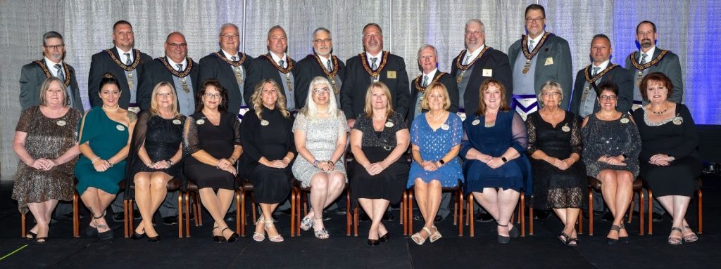Large group portrait of Ohio Masonic officers in formal regalia standing behind their spouses and partners seated in evening attire on a stage during an Annual Communication.