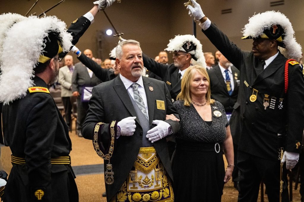 A couple walks arm-in-arm beneath raised swords held by Knights in full dress uniforms with plumed hats during the installation ceremony.