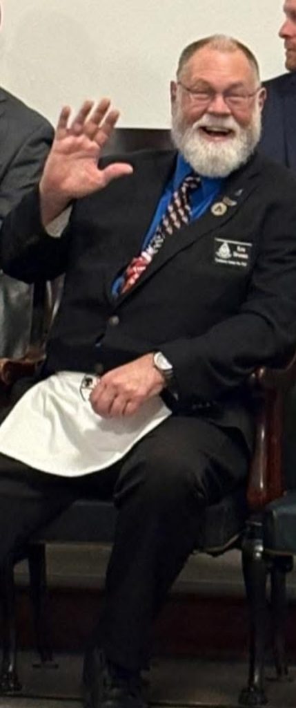 A seated Freemason in formal attire smiling and raising his hand during a lodge gathering.