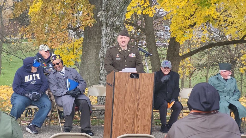 A man dressed in a United States Army uniform, speaks to a crowd outdoors below a tree with yellow leaves, and behind a brown wooden podium.